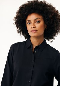 Černá košile na knoflíky s límečkem, která má hladkou texturu a dlouhé rukávy. Tkanina se zdá být lehká a volně strukturovaná.