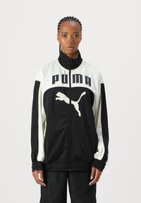 PUMA sportlik jakk, millel on must keha ja valged aktsendid, kõrge krae, täiskraan ja suur logo rinna peal. Elastsed mansetid.