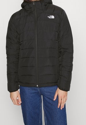 Persona vestida con una chaqueta con capucha acolchada negra de The North Face, cerrada con cremallera, y jeans de mezclilla azul, de pie contra un fondo liso.