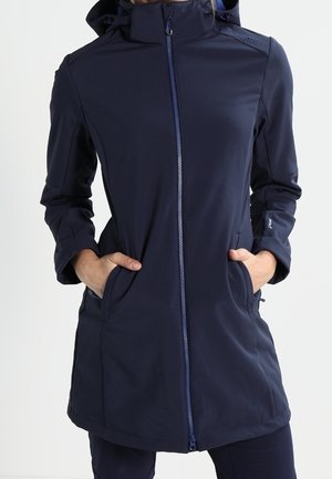 Vrouw draagt een lange marineblauwe jas met ritssluiting, met handen in de zijzakken, voorzien van een hoge kraag en een capuchon.