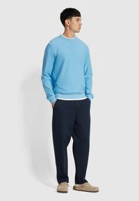 Maglione azzurro chiaro con collo rotondo, polsini a coste e un piccolo logo, abbinato a pantaloni blu scuro e scarpe beige senza lacci. Sfondo semplice.