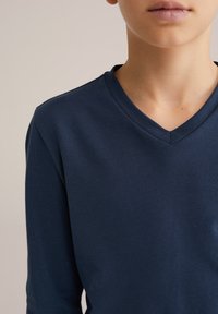 Tengeri kék hosszú ujjú V-nyakú póló puha, rugalmas anyagból. Sima tapintású, a nyakkivágásnál bordázott részlettel, szorosan illeszkedik.