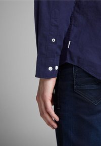 Camicia a maniche lunghe in cotone blu navy con bottoni bianchi sui polsini, dettaglio laterale con spacco e tessuto liscio, abbinata a jeans denim scuro.
