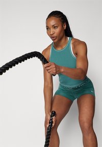 Idrettsbekledning inkluderer en teal rippet tanktopp med hvite detaljer og matchende shorts. Personen griper en tykk, svart treningsrep.