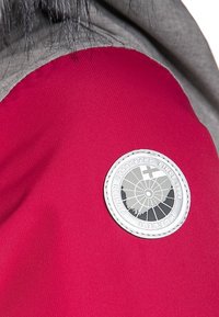 Szczegół czerwonej i szarej kurtki z okrągłą, srebrną naszywką z okrągłym wzorem i krzyżem. Gładka tekstura materiału, podkreślone przeszycia.