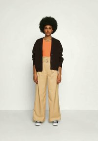 Cardigã castanho-escuro, top laranja, calças bejes de cintura alta, cinto bege e ténis pretos com solas brancas; malha texturizada e design de perna ampla.