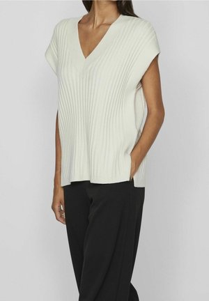 Kvinde iført hvid ærmeløs ribstrikket V-hals sweater og sorte bukser, stående med den ene hånd i lommen mod en ensfarvet baggrund.
