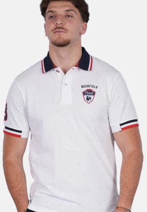 Homme portant un polo blanc avec un col marine et des bord-côtes de manches rayés rouge, blanc et bleu, avec le logo "Ruckfield French" sur la poitrine.