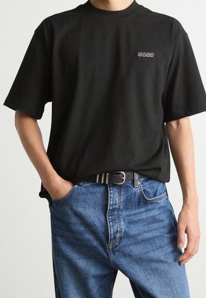 Person iført en sort T-shirt med "BOSS" logo på brystet, delvist stikket ned i blå jeans med sort bælte, den ene hånd i lommen.