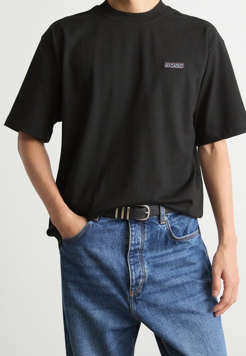 Personne portant un T-shirt noir avec le logo "BOSS" sur la poitrine, partiellement rentré dans un jean bleu avec une ceinture noire, une main dans la poche.