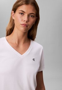 T-shirt bianca in cotone con scollo a V e maniche corte, con un piccolo logo nero "CK" sull'area del petto sinistro.