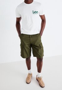 Camiseta de algodón blanca con el logo verde de "Lee", combinada con pantalones cortos cargo verde olivo que tienen bolsillos laterales, llevada con zapatos beige y calcetines blancos.