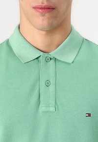 Svetlozelená polo košela z textúrovanej tkaniny, s golierom, dvoma gombíkmi a malým vyšitým logom s červenými, bielymi a modrými akcentmi.