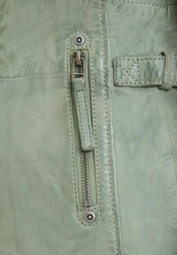 Lichtgroen leer met een zichtbare ritsdetail, zilverkleurige hardware en contrasterende stiksels langs de randen. Gladde textuur.