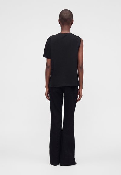 Personne debout de dos à la caméra portant un haut asymétrique noir à manches courtes et un pantalon évasé noir sur un fond clair uni.