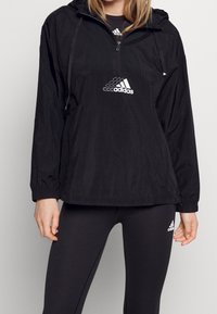 Zwarte windbreaker gemaakt van lichtgewicht stof met een capuchon, uitgerust met een halve rits aan de voorkant en een wit Adidas-logo op de borst.