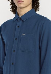 Chemise longue à manches bleues avec un col boutonné, poche avant et boutons noirs, fabriquée en tissu texturé. Étiquette de marque visible.