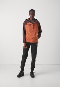 Oransje og brunt hetteanorak med en frontlomme, elastiske mansjetter, kombinert med svarte smalere bukser og svarte turstøvler.