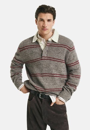 Junger Mann in einem grauen Strickpullover mit roten Streifen über einem cremefarbenen Hemd mit Kragen, schwarzem Gürtel und dunklen Hosen, der mit den Händen in den Taschen steht.