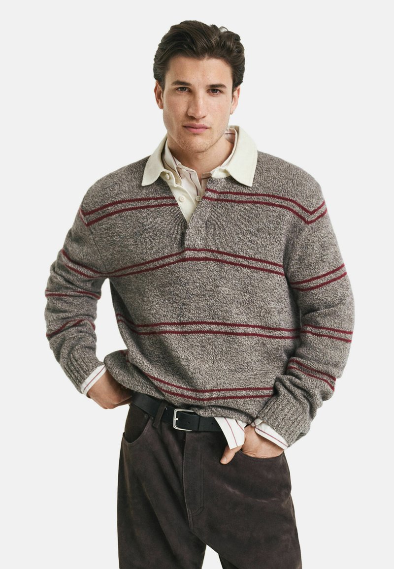 Un jeune homme portant un pull en tricot gris avec des rayures rouges sur une chemise crème à col, une ceinture noire et un pantalon sombre, debout les mains dans les poches.