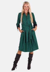 Grünes kariertes Kleid mit langen Ärmeln, schwarzen Akzenten, Bindegürtel und Taschen. Getragen mit knielangen schwarzen Stiefeln. Glatter, strukturierter Stoff.