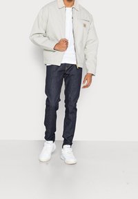 Chaqueta bomber gris claro con un cuello beige, camiseta blanca de cuello redondo, vaqueros azul oscuro y zapatillas blancas. Detalle de bolsillo en el pecho con cremallera.