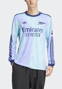 Lyseblå langærmet fodboldtrøje med gradientdesign, marineblå detaljer, Adidas-logo og teksten "Emirates Fly Better" på forsiden.