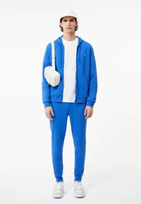 Haut à capuche bleu avec poches sur le devant, associé à un pantalon de survêtement assorti. T-shirt blanc en dessous et sac bandoulière blanc. Des baskets complètent le look.