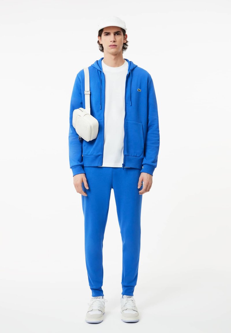 Felpa blu con tasche frontali, abbinata a pantaloni della tuta coordinati. Maglietta bianca sotto e borsa a tracolla bianca. Sneakers completano il look.