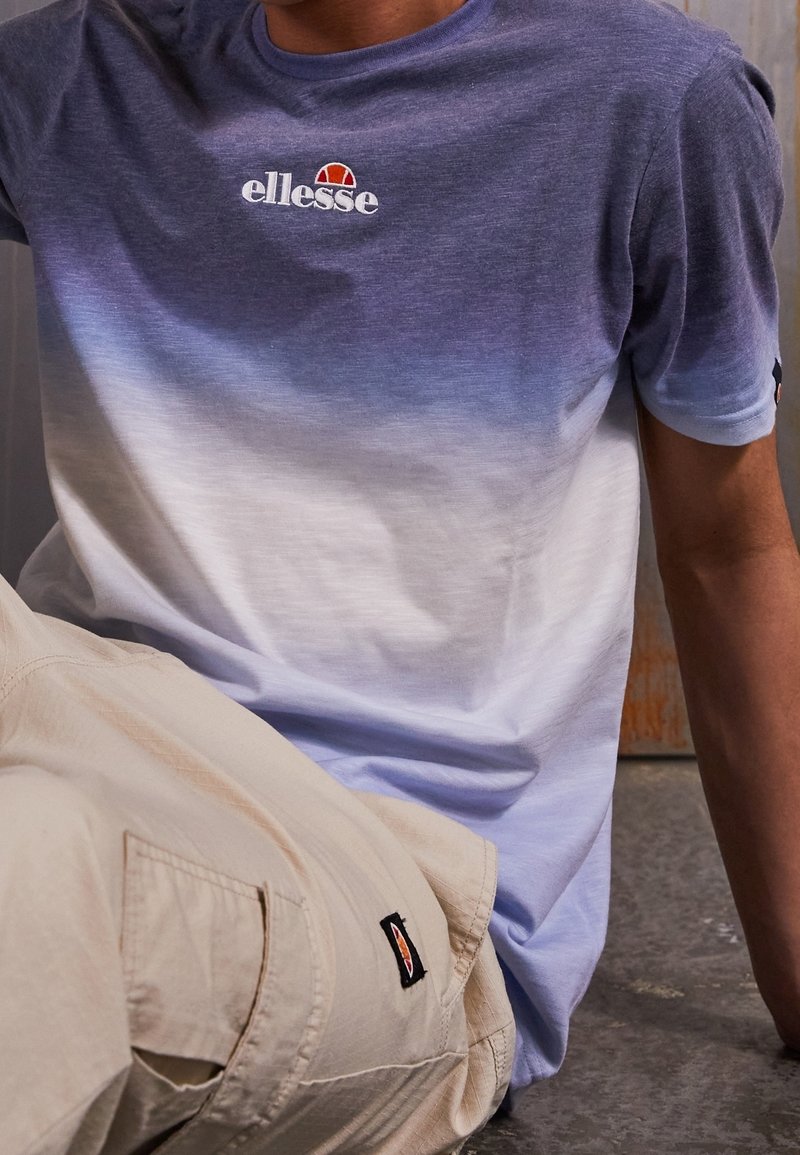 Personne portant un t-shirt Ellesse bleu et blanc en dégradé et un pantalon beige clair, assise avec le bras posé au sol. Les logos Ellesse sont visibles.