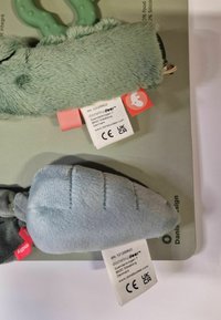 Two plush toys: a soft green animal and a light blue carrot. Both have fabric tags with care instructions prominently displayed.