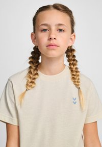 Beige bomulds t-shirt med rund hals og korte ærmer, med et lyst blåt pil-design på venstre bryst. Glat tekstur.