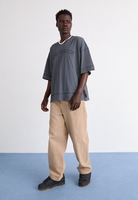 Homme debout sur un tapis bleu, portant un t-shirt gris foncé surdimensionné, un pantalon beige, des baskets noires et un collier blanc en perles, regardant vers la droite.