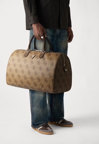 Sac de voyage à motifs marron avec des poignées en cuir, doté d'une fermeture éclair et d'accents en métal. Porté avec un jean en denim bleu délavé.