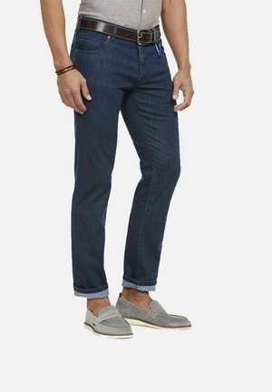 Man met opgerolde donkerblauwe jeans, grijze loafers, een bruine leren riem en een lichtgrijs, in gestoken overhemd, staat met ontspannen handen.