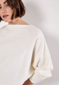 Witte, gestructureerde top met een ontspannen pasvorm, boothals en volumineuze mouwen met subtiele ribbels.