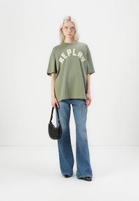 Groen oversized t-shirt met de tekst "REPLAY" in crème, gecombineerd met lichtblauwe flared jeans. Gemaakt af met een zwarte geperforeerde tas.