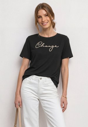 Femme souriante portant un t-shirt noir avec le texte "Change" et un pantalon blanc, tenant la lanière d'un sac à main beige d'une main.