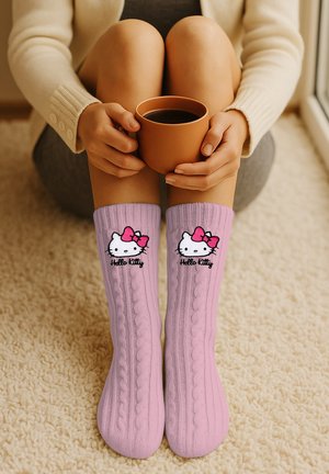 Person sitting on carpeted floor holding a brown cup, wearing pink Hello Kitty socks with cable knit pattern.