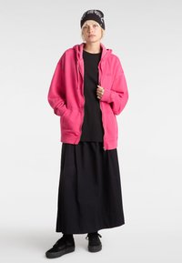 Pink zip-up hoodie with two front pockets, layered over a black T-shirt and a long black skirt, paired with black shoes and a black beanie.