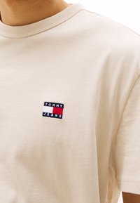 Vaaleanoranssi lyhythihainen T-paita, joka on valmistettu pehmeästä puuvillasta. Edessä on pieni, brodeerattu tummansininen, punainen ja valkoinen suorakulmainen logo rintamuksessa.