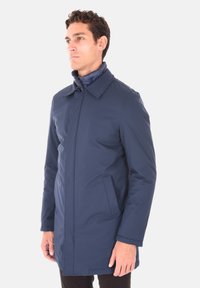 Cappotto impermeabile blu navy con colletto, frontale con zip e due tasche laterali. Presenta una texture liscia e una vestibilità strutturata.