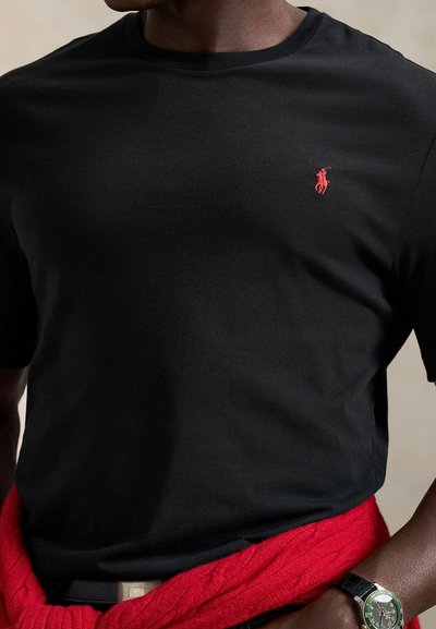 Černé bavlněné tričko s kulatým výstřihem, na hrudi malý červený vyšívaný logo polo. Červený svetr uvázaný v pase.