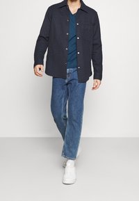 Giacca trapuntata navy con chiusura a bottone, indossata sopra una maglietta teal a scollo a V, abbinata a jeans azzurri e sneakers bianche.