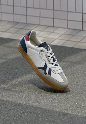 Weißer Sneaker mit dunkelblauen und roten Akzenten, beiger Sohle und weißen Schnürsenkeln, auf grauem Pflaster nahe einer gefliesten Wand ruhend.