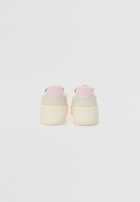 Back view of sneakers featuring a soft beige body, pink accents at the heel, and a textured rubber sole. Smooth material with slight sheen.