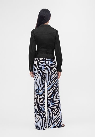 Femme aux longs cheveux foncés portant une veste noire cintrée et un pantalon large avec des rayures zébrées abstraites noires, blanches et bleues, debout de dos.