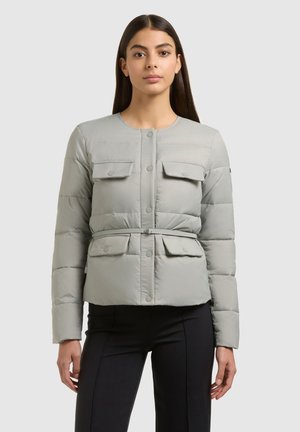 Woman with long dark hair wearing light grey belted quilted jacket with four flap pockets, paired with black fitted pants, against plain background.