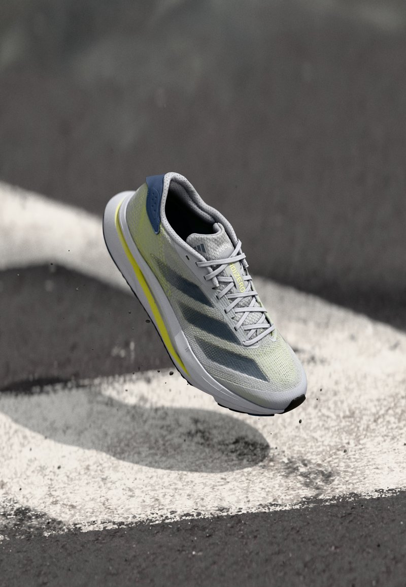 Chaussure de course légère avec une tige en mesh gris, un accent jaune, un contrefort bleu et une semelle blanche. Présente un design à trois bandes et un système de laçage.