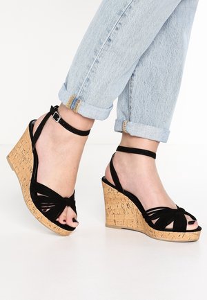 Black wedge sandals with cork platform, featuring a knotted strap design and adjustable ankle strap. Worn with rolled-up denim jeans.